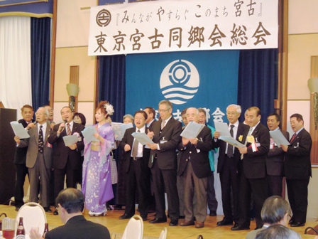 ”『みんながやすらぐこのまち宮古』東京宮古同郷会総会”と書かれた舞台看板が掲げられ、真ん中で紫色の着物を着た女性とそのまわりにスーツを着た男性達が紙を持ち歌を歌っている写真
