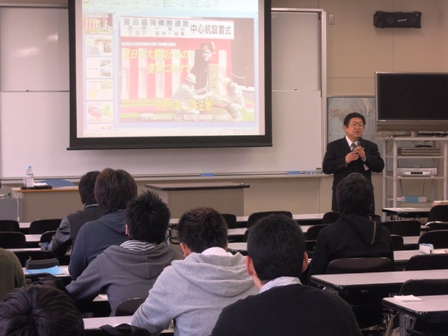 ホワイトボードの前に設置されたスクリーンに写し出される資料をもとに講演をする市長と、参加者の方々の写真
