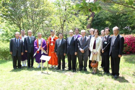 青々とした木々の中に植樹した桜の前で、市長と関係者13名が横2列に並んでいる写真