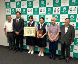 バックパネルの前で賞状と盾を持った制服を着た女子中学生2名と、市長を含む4名の男性が横一列に並び記念撮影をしている写真