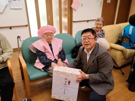 施設の敬老会でピンクの頭巾とちゃんちゃんこを着て、椅子に座っている横田ミネさんに 市長が市からの記念品を渡している写真