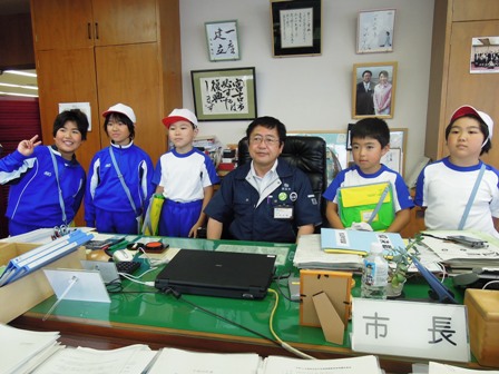 市長室で席に座る市長の両側に、体操服姿の小学生5人が並んでいる写真