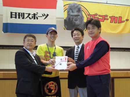 日刊スポーツとゴーゴーカレーと書かれた旗が壁に飾られており、市長と工藤公康さん、関係者4人が目録に手を添えている写真