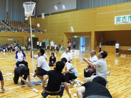 広い体育館の中で、長い棒の先端に網のかごをつけた棒を下から男性が支えており、周りの参加者が網かごに向けて白い柔らかい玉を投げ入れている写真