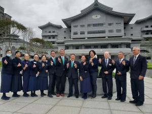 曇り空の中グレーの外壁の關渡靜思堂(カントセイシドウ)が奥に見える屋外で、関係者の方々と1列に並んで右こぶしをにぎり記念撮影をしている写真