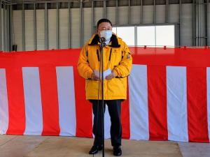紅白幕が飾られた安全祈願祭と除雪出動式の会場で、黄色のジャンパーを着用した市長がマイクスタンドの前で挨拶を行っている様子の写真