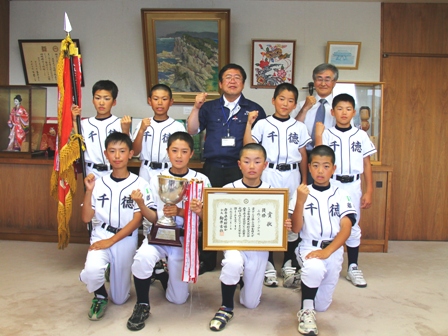 千徳と書かれたユニフォームを着た男子生徒達が優勝旗や優勝カップ、賞状を持ち市長とガッツポーズをして記念撮影している写真