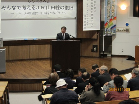 演題の上にのっているスタンドマイクの前で話をしている市長と長テーブルの前に着席して話をきいている参加者の写真