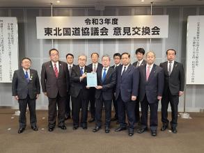 「令和3年度東北国道協議会 意見交換会」と書かれた横断幕が掲げられた室内で、国土交通省の村山一弥道路局長と代表の男性が要望書を一緒に持ち、市長を含め9名の関係者の方々が2列に並んでいる写真