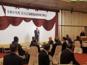 「令和6年度 宮古広域圏地域情報交歓会」と書かれた横断幕が吊るされた舞台上でスタンドマイクに向かい話をする様子の市長と、円卓になったテーブルの席につき話を聞く様子の参加者たちの写真