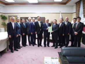国旗が置かれた室内で、鈴木俊一財務大臣と市長が要望書を一緒に持ち10名の関係者の方々が横一列に並んでいる写真