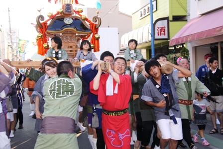 神輿の先頭に赤い浴衣をきた市長と参加者たちが、商店街の道を子供達をのせた神輿を担ぎながら歩いている写真