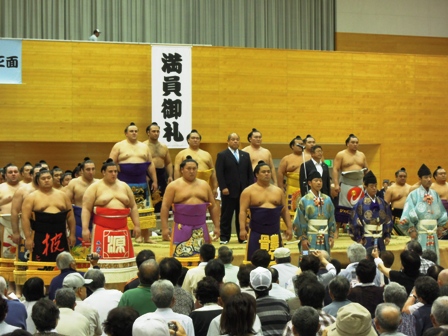 「満員御礼」と書かれた垂れ幕の前の土俵や土俵の回りに力士や行司の方々が集まり真ん中にスーツ姿の男性立っているのを撮影している写真