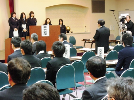 前方に発表者席と書かれた演説台に5人の女子高校生が横一列になり、向かい合わせで市長がマイクを持ち話している後姿の写真