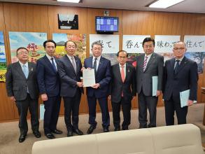 壁に大きなポスターがきれいに並んで掲示されている室内で、復興庁の富樫博之副大臣と代表の男性が要望書を一緒に持ち、市長を含め5名の関係者の方々が横一列に並んでいる写真