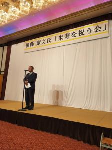 「後藤康文氏「米寿を祝う会」」と書かれた横断幕が吊るされた舞台上で、スタンドマイクに向かい話をする様子の市長の写真