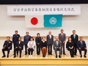 「宮古市自衛官募集相談員委嘱状交付式」と書かれた横断幕と日本国旗、宮古市の市町村旗が吊るされた舞台上で、前列に椅子に座る7名の男女、後方に横一列に立つ6名の男女が写っている記念写真