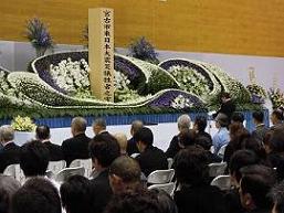 白い花で飾られた祭壇の前で、市長が式辞を述べているのを多くの参加者の方々が座って聞いている写真