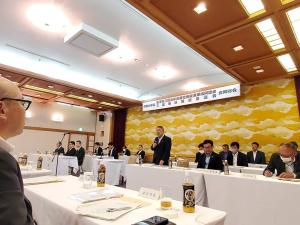 白いテーブルクロスが敷かれた机が向かい合うように並べられた会場の中心で、マイクを右手にマイクを持ち立って話をしている様子の市長の写真