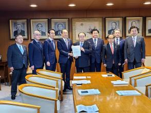 一人掛けの大きな椅子と横長の机が設置された室内で、自由民主党の田中和徳幹事長代理と代表の男性が要望書を一緒に持ち、市長を含め7名の関係者の方々が横一列に並んでいる写真