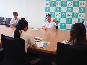 広い会議室で、手ぶりを交えて話をする市長と、対面して話を聞いている青山学院高等部の女子生徒を後方から撮影した写真