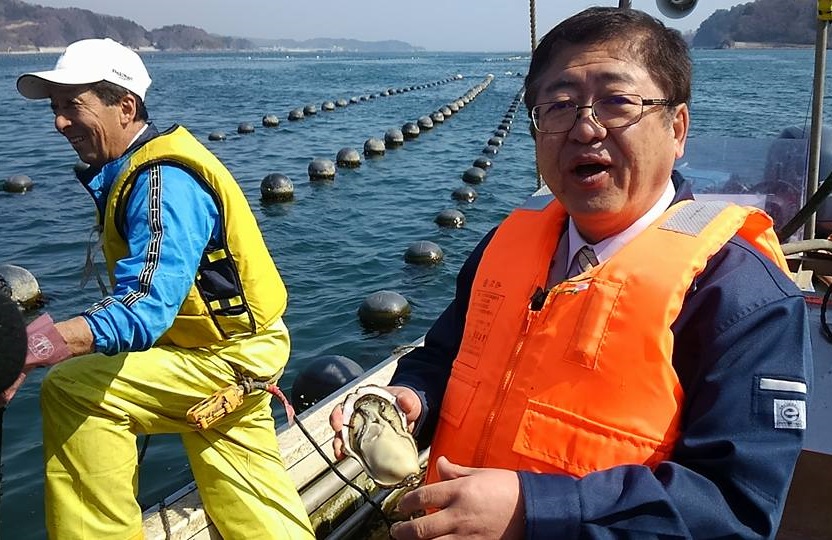 海の上で、ライフジャケットを着た市長が手のひらほどのカキを手に持っている写真