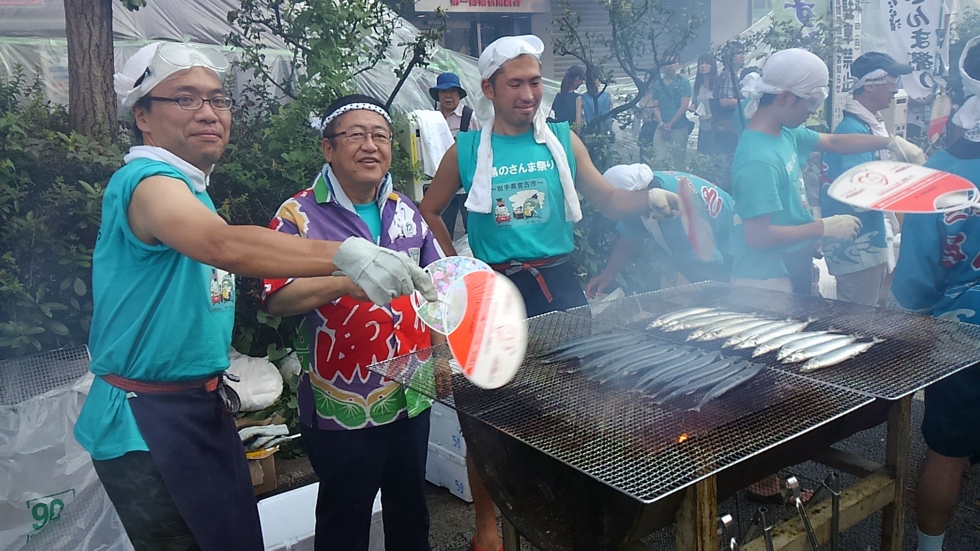 市長と関係者が、網の上で焼かれているさんまをうちわで仰いでいる写真