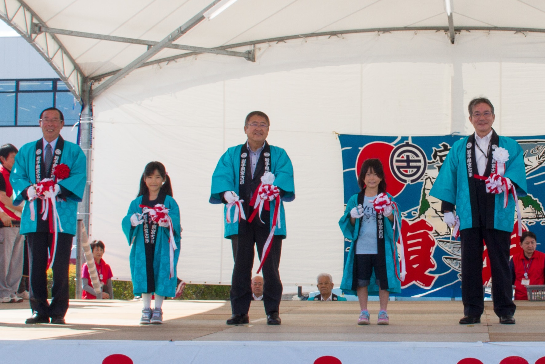 はっぴを着た市長と子供と関係者の5名が、ステージ上でテープカットをしている写真