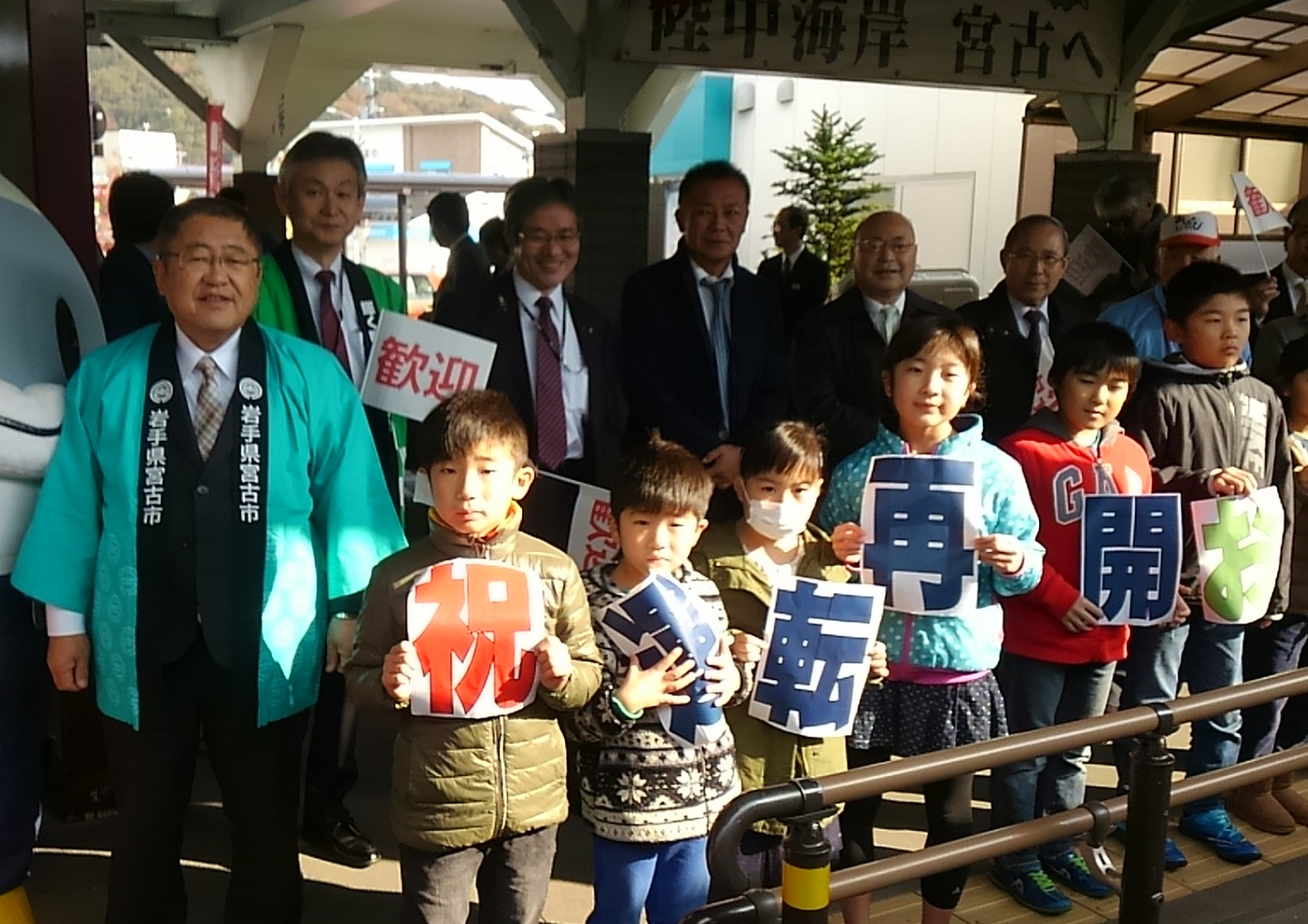 駅の構内で、はっぴを着た市長と、「祝 運転再開」の文字パネルを持つ子供達と関係者の写真