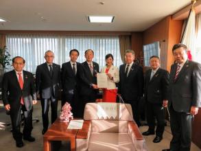 国旗が置かれ応接セットが設置された室内で、経済産業省の吉川ゆうみ大臣政務官と代表の男性が要望書を一緒に持ち、市長を含め6名の関係者の方々が横一列に並んでいる写真