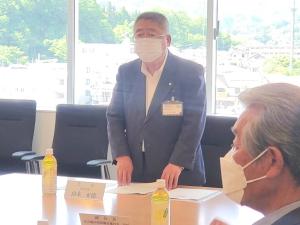 名前札とペットボトルのお茶が置かれたテーブルと黒い椅子の間に立ち話をしている様子の市長の写真
