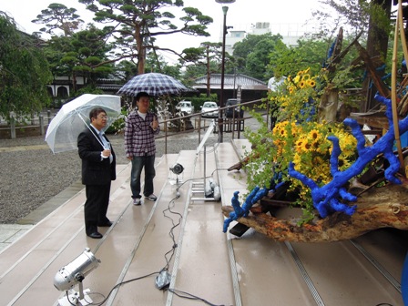 護国寺の階段に置かれた太い木の枝のような震災がれきに、ひまわりの花や緑が添えられて展示されており、それを眺めている市長と関係者の写真