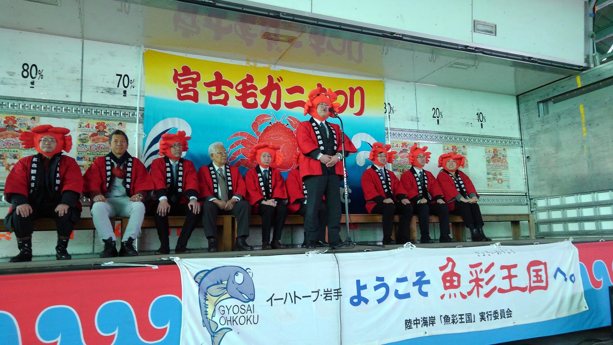 「宮古毛ガニ祭り」と書かれた幕が設置された特設ステージ上でカニの被り物をした市長がスタンドマイクで話をし、後ろにカニの被り物をした関係者が並んで座っている写真