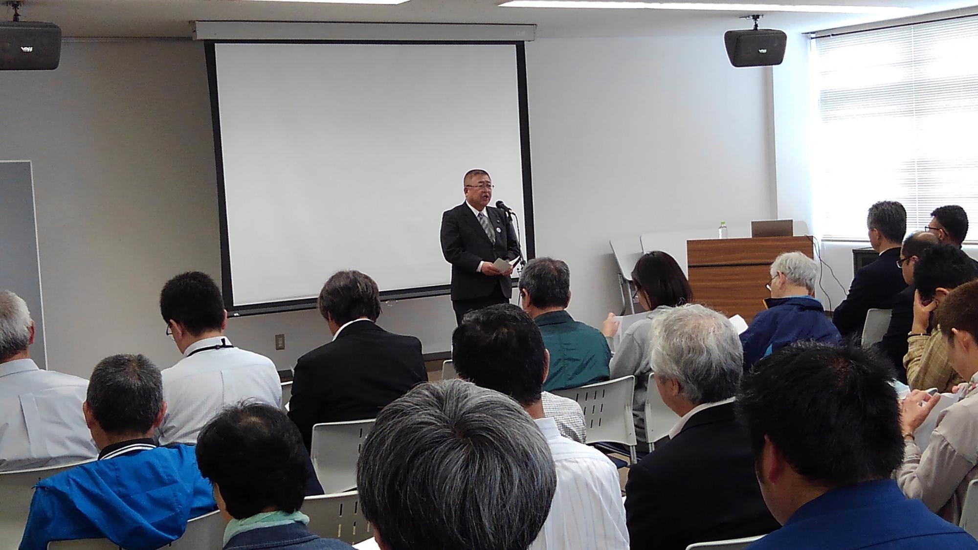 天井から吊るされたスクリーンの前で市長が話をしているのを聞く参加者たちの写真