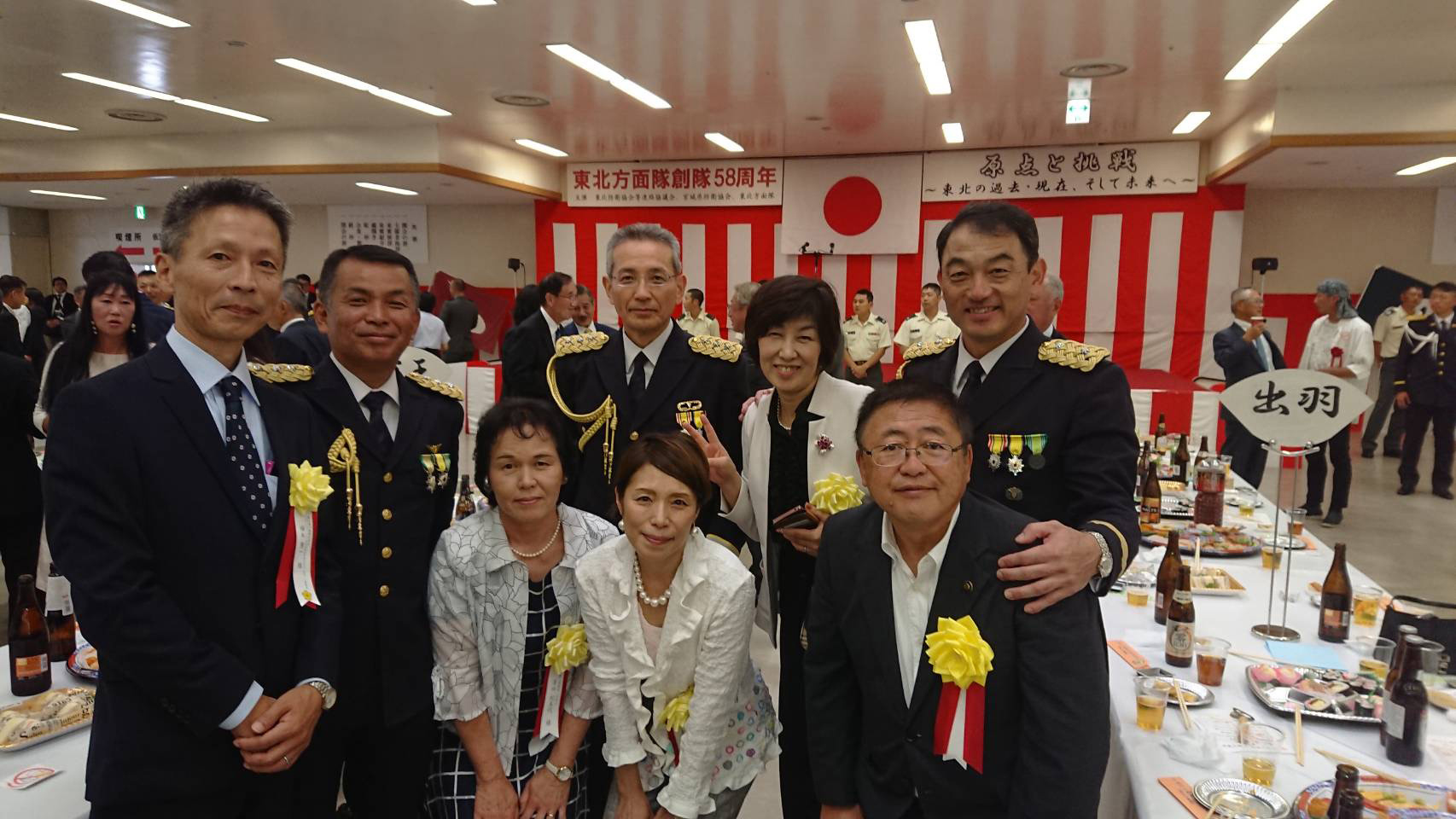 料理や飲み物が置かれた長机の横で胸章を胸につけた市長と関係者たちが記念撮影している写真