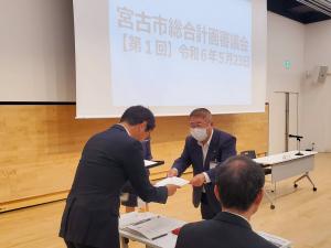画面が映されたスクリーンの横で、関係者の男性に紙を両手で手渡す様子の市長の写真