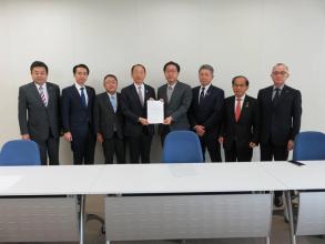 一脚ずつ椅子がセットされた長机が並んだ室内で、文部科学省の池田貴城研究振興局長と代表の男性が要望書を一緒に持ち、市長を含め6名の関係者の方々が横一列に並んでいる写真
