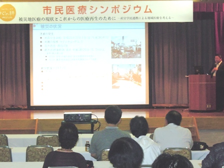 ステージ上に「市民医療シンポジウム」の横断幕があり、中央にスクリーンで映し出される資料を見ながら説明をする市長と、椅子に座り耳を傾けて話を聞く参加者の写真