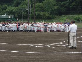 新里山村広場のグランドで、少年野球チームがプラカードや優勝旗を手に持ち、チームごとに整列しており、向かい合って挨拶をする市長の写真
