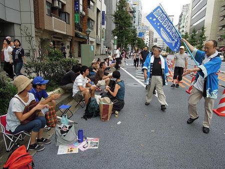 道路にアウトドアチェアに座っている参加者たちの前を青色の半被を着た市長が歩いている写真