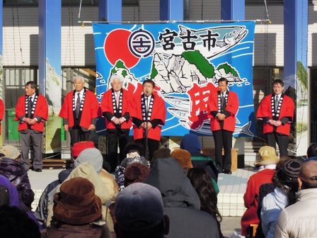 「宮古市」の文字と鮭の絵が描かれた横断幕の前で赤い半被をきた関係者達と市長がマイクを使い挨拶している写真