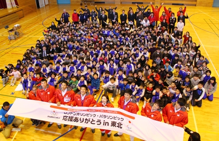 広い体育館に「応援ありがとう in 東北」の横断幕を手に持つオリンピック選手8名と、その後ろに集合して座る大勢の小学生や関係者達がポーズを取っている写真