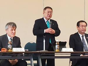 マイクを持ちながら起立して挨拶をしている市長と関係者2名が着席して聞いている写真