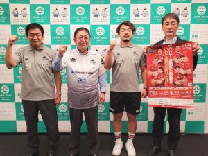 ラグビーチーム釜石シーウェイブスの坂下功正総監督と、小野航大キャプテン、市長とポスターを持った男性がバックパネルの前で記念撮影をしている写真