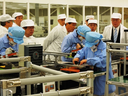 機械が置かれた工場内で白色と青色の作業着を着た人たちが作業しているのを市長が見学している写真