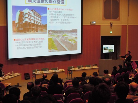 会場のホールのスクリーンに資料が映し出されており、横の演壇で説明する市長と、話に耳を傾ける参加者の写真
