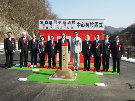 「宮古盛岡横断道路中心杭設置式」と書かれた横断幕の下に紅白幕があるステージと中心杭の間にスーツを着た関係者たちがが並び記念撮影している写真