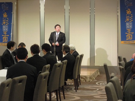 ステージでマイクスタンドの前に立ち話をする市長と、座席に座り話を聞く関係者達の写真