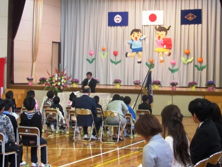 小学校のステージ壁面にランドセルを背負った男の子と女の子の切り絵が色鮮やかに飾られており、演壇で祝辞を述べる市長の話を新入生や在校生が椅子に座って聞いている写真
