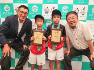 首からメダルをかけ賞状を持った久保尻翔真さん、久保尻隼翔さんが真ん中に立ち、市長と男性がその両脇で久保尻さんと目線をあわせるようにかがんでいる記念写真